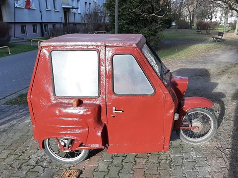 Red three-wheeler with a boxy steel cabin and two windows, paint peeling at the door, parked on paving.