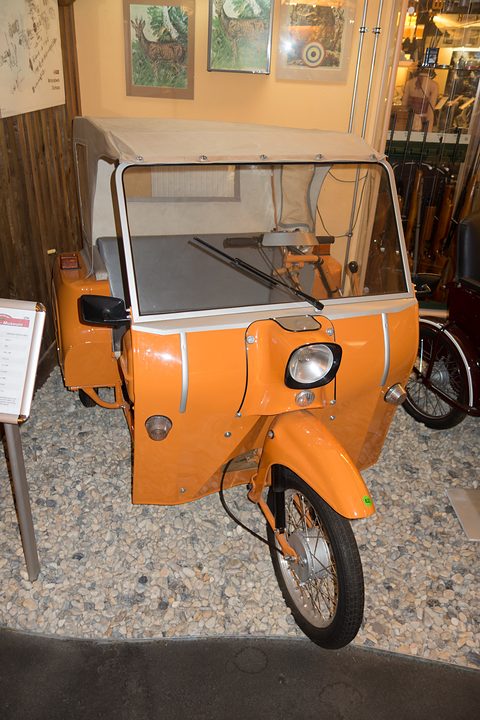 Orange three-wheeler with a single front wheel, a bare hood frame and a windscreen, on gravel in a small museum. Opens a larger view