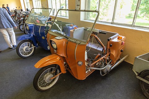 Orange three-wheeler without a hood, its windscreen and grey seat visible, in a hall lined with blue and red scooters. Opens a larger view