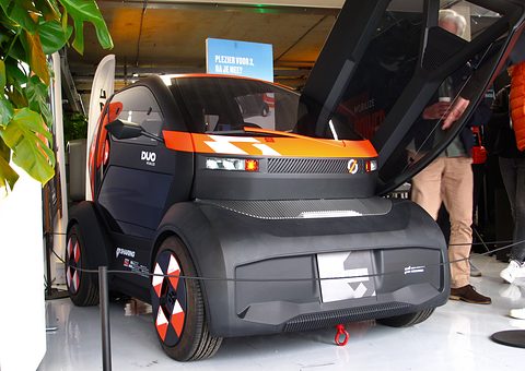 Dark grey and orange tiny two-seater with its door raised on a show floor, visitors standing beside it.