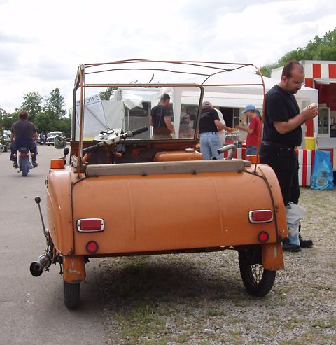 Rear of a tan three-wheeler with a rounded tail, two red lamps and a bare tubular hood frame, people at a show behind. Opens a larger view