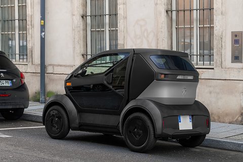 Rear of a black and grey tiny two-seater with a light-strip tail, parked behind a dark hatchback. Opens a larger view