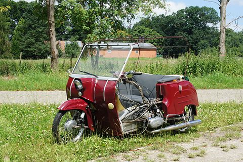 Dark red three-wheeler with a bare windscreen and no hood, standing on a grass verge by a road and trees. Opens a larger view