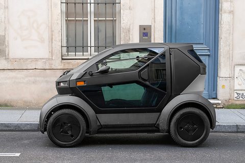 Side view of a short black and grey two-seater with a large door and big wheels, parked by a stone house. Opens a larger view