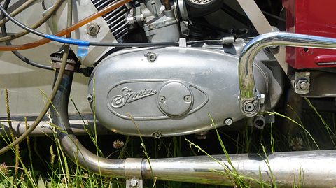 Close view of a silver single-cylinder engine with a Simson script on its cover and a chrome exhaust, grass below. Opens a larger view