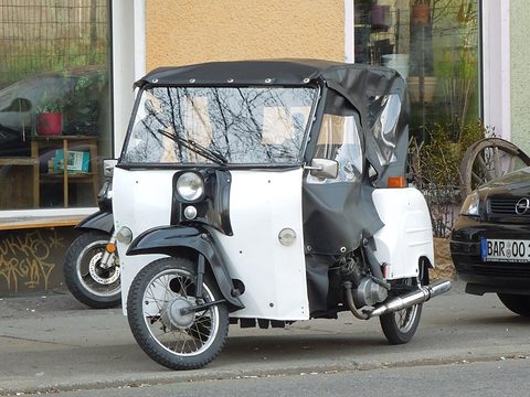 White three-wheeled invalid carriage with black mudguards and a black hood with clear windows, at a kerb. Opens a larger view