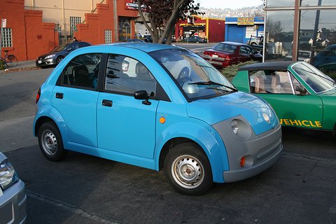 Light-blue rounded four-door small car with grey bumpers parked in a street, a green open car behind. Opens a larger view