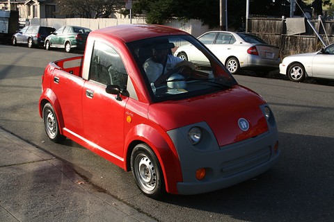 Red small two-door pickup with a grey nose and round lamps on a street, its driver visible through the glass.