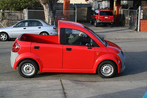 Side view of a red small pickup with a short bed and grey bumper on a sunlit street, a man at the wheel. Opens a larger view