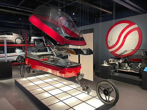 Red and white three-wheeler body hanging on wires over its wheels and chassis on a lit floor, more cars behind.
