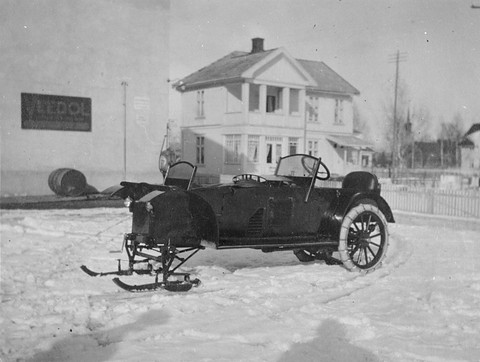 A dark open car standing on snow, skis under its front and chained spoked wheels at the back, a white house behind. Opens a larger view