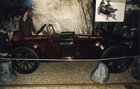 A long dark-red open car with wooden wheels beside a mannequin in a coat, painted snowy trees and a period photograph behind. Opens a larger view