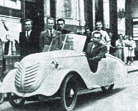 A pale streamlined open two-seater with a louvred nose and enclosed wheels, a man at the wheel and several men behind.