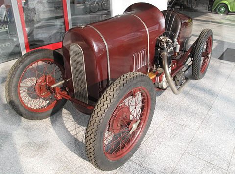 A small maroon racing car with a mesh radiator cowl, red wire wheels and an air-cooled single-cylinder engine at its side.