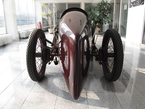 Rear view of a slim maroon racing car whose body tapers to a point, spoked wheels on either side on a polished floor. Opens a larger view