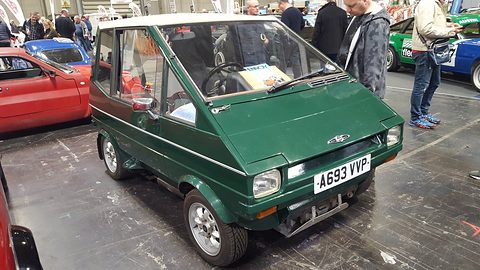 Dark green upright city car with rectangular headlamps on a show floor, visitors around it. Opens a larger view