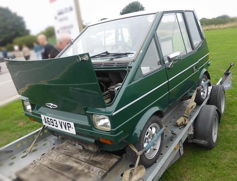 Green boxy car strapped to a trailer on grass, its front-hinged bonnet lifted. Opens a larger view