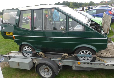 Dark green upright car side-on on a flatbed trailer at an outdoor show. Opens a larger view
