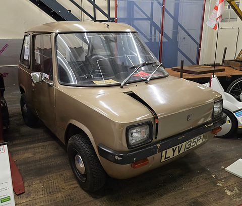 Beige upright electric car on a wooden floor among museum exhibits. Opens a larger view