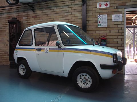 White electric car with blue and yellow stripes parked in front of a brick wall with safety signs.
