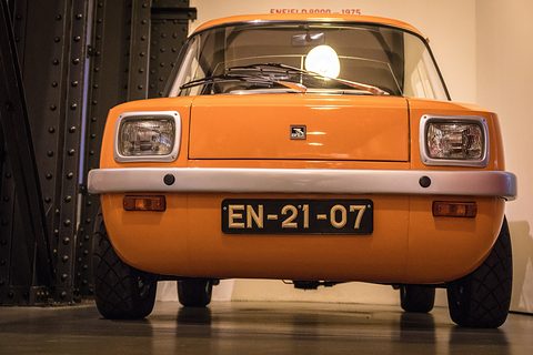 Orange box-shaped electric car facing the camera, a lamp glowing behind it in a gallery. Opens a larger view