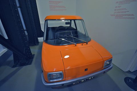 Orange electric car seen from above at the front quarter beside dark steel girders. Opens a larger view