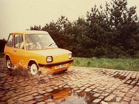 Orange car splashing across wet cobbles in front of trees. Opens a larger view