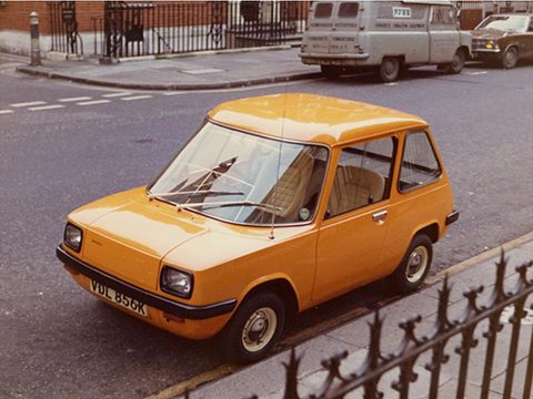 Orange electric car at a kerb with iron railings in the foreground and a van behind. Opens a larger view