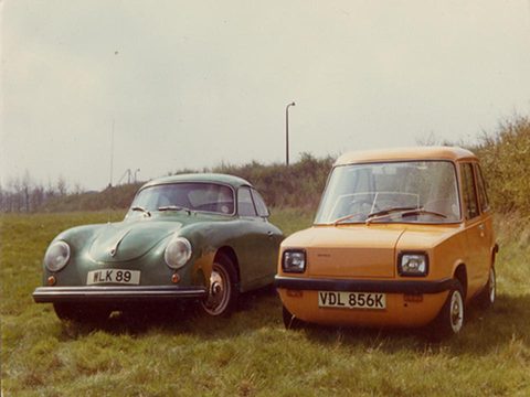Orange box-shaped car next to a dark green Porsche coupé on grass. Opens a larger view