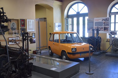Small car in a museum hall with arched windows, old machinery around it.