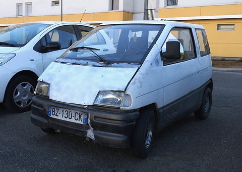 Front of a tired white small car with a dented bonnet and black bumper, parked beside a modern white hatchback.