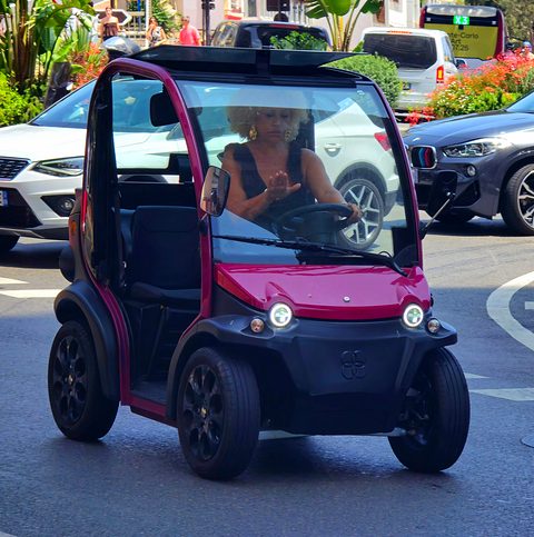 Front of a narrow red and black two-seat microcar with round lamps and an open side, driven along a street. Opens a larger view