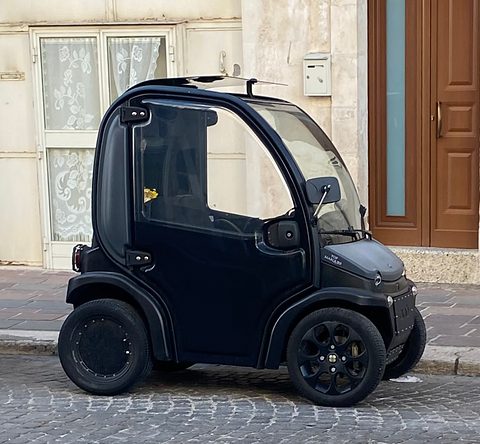 Side view of a black tall, narrow two-seat microcar on a cobbled street by a lace-curtained window. Opens a larger view