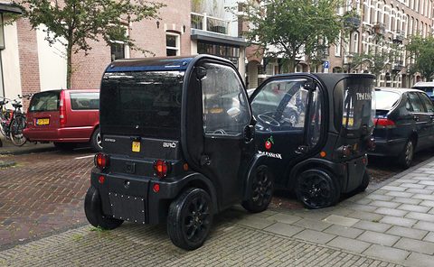 Two tall, narrow black two-seat microcars with large wheels parked on a pavement in front of brick houses. Opens a larger view