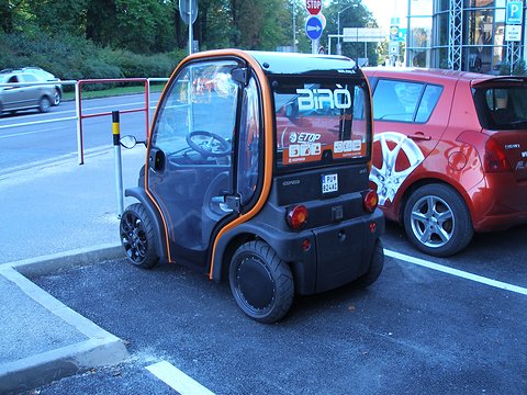 Black and orange tall two-seat microcar with BIRÒ lettering on its rear glass, parked next to a red hatchback. Opens a larger view