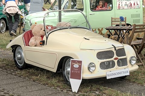 Cream open two-seater with a KLEINSCHNITTGER plate on grass, a teddy bear aboard and a pale green van behind. Opens a larger view