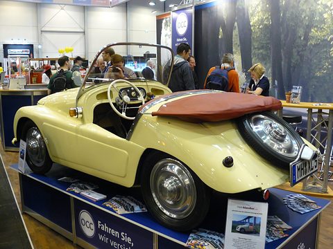 Cream open two-seater with a tan tonneau and a spare wheel on its tail, on a blue show plinth. Opens a larger view