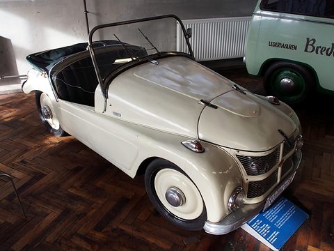 Cream open two-seater with full-width grille and a blue information board on a parquet floor. Opens a larger view