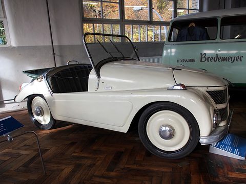 Cream open two-seater in side view on parquet, a Bredemeyer leather-goods van beside it. Opens a larger view