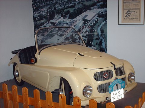 Cream open two-seater with a mesh grille and rally plaques behind a wooden fence, an aerial photograph behind. Opens a larger view