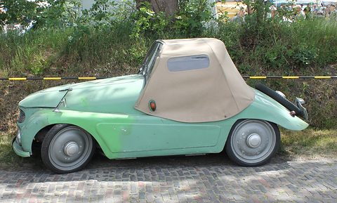 Faded green open two-seater with a beige fabric hood raised, parked on cobbles before a hedge.