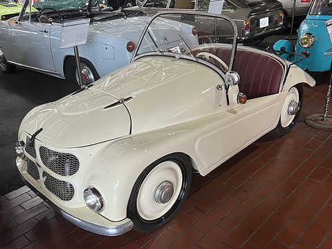 Cream open two-seater with maroon seats on a red tiled floor among other small cars. Opens a larger view