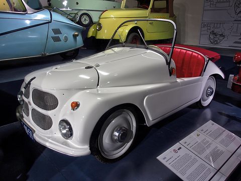 White open two-seater with red seats on a dark display beside a blue bubble car and a yellow microcar. Opens a larger view