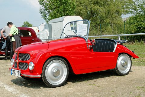 Red open two-seater seen from the front left on gravel, visitors and a caravan behind. Opens a larger view