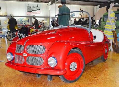 Bright red open two-seater with red wheels on an orange floor, period-dressed mannequins behind. Opens a larger view