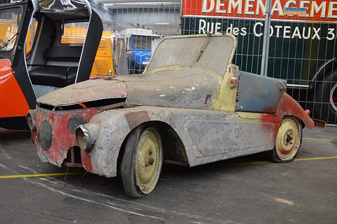 Battered open two-seater with peeling red, grey and cream paint on a hall floor, a bubble car beside it.