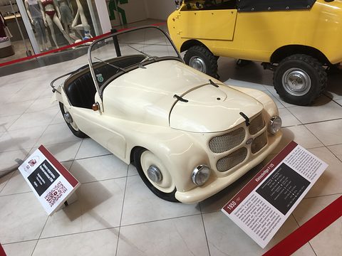 Cream open two-seater on a white tiled floor between two information boards, a yellow microcar behind. Opens a larger view