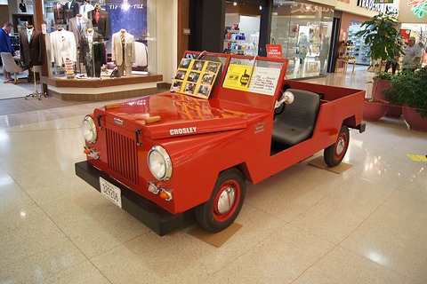 Small red Jeep-like utility with a vertical-slot grille, round headlamps and a folded screen, on a shiny shopping-mall floor. Opens a larger view