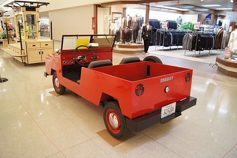 Rear quarter of a small red utility with a square load box lettered CROSLEY, two seats and small wheels, in a shopping mall. Opens a larger view