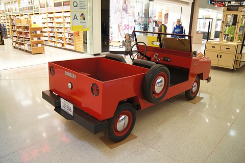 Small red utility from the rear right, a spare wheel mounted on its side, two seats and a load box, before shop fronts. Opens a larger view